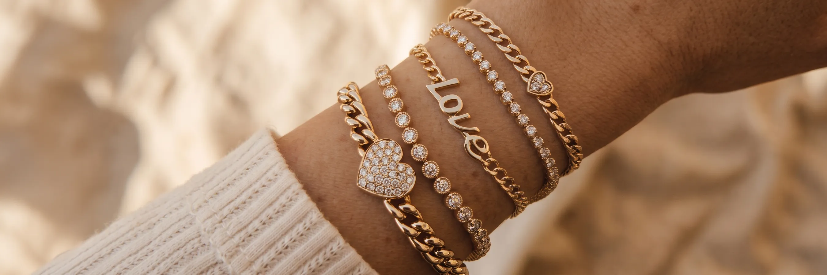 A wrist adorned with multiple gold bracelets featuring hearts, diamonds, and the word