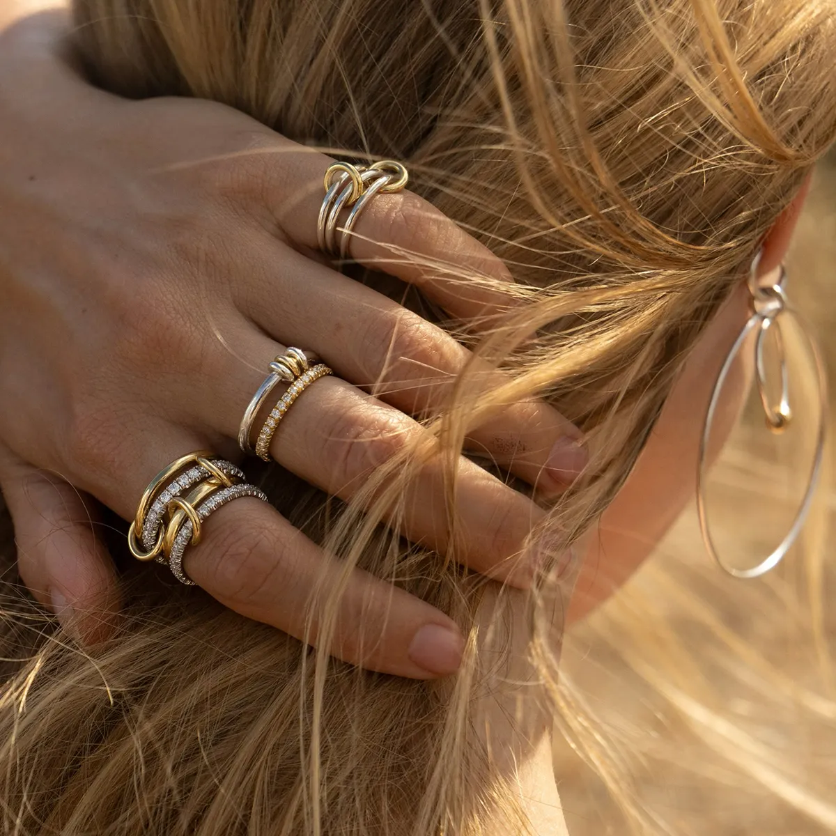 A hand with multiple gold and silver rings holding blonde hair, with a silver hoop earring visible.