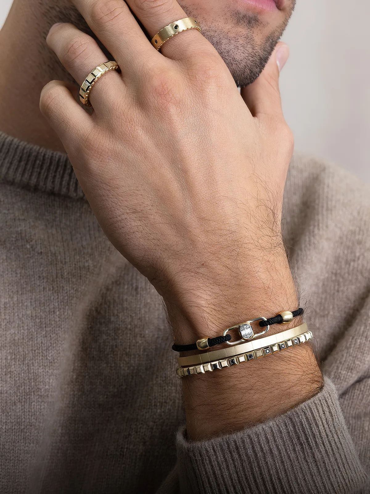 A person wearing gold rings and bracelets, with a hand on their face, dressed in a beige sweater.