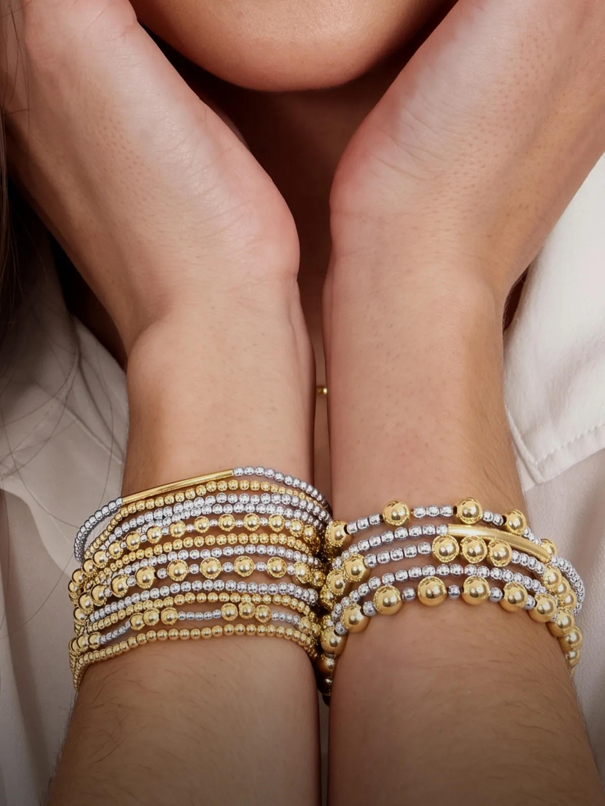 Stacked gold and silver bead bracelets on wrists, hands near face.