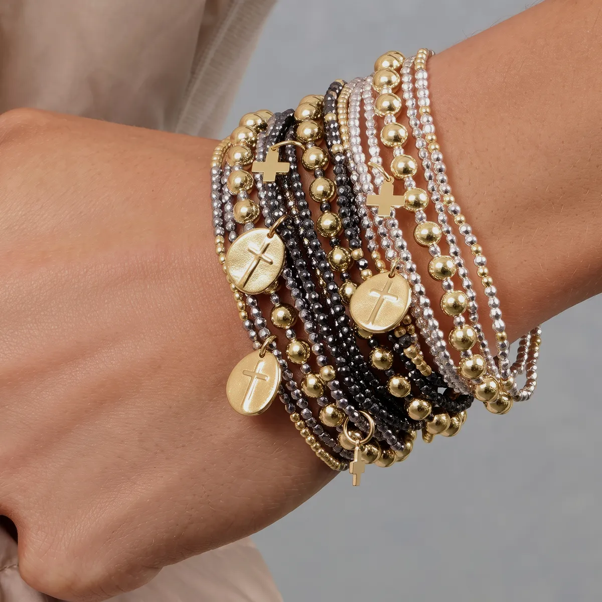Multiple gold and silver beaded bracelets with cross and disc charms on a wrist.