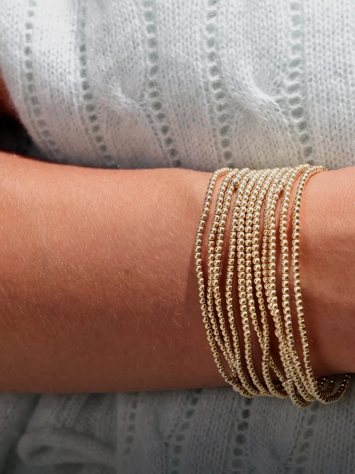 A wrist adorned with multiple thin gold beaded bracelets against a textured white fabric background.