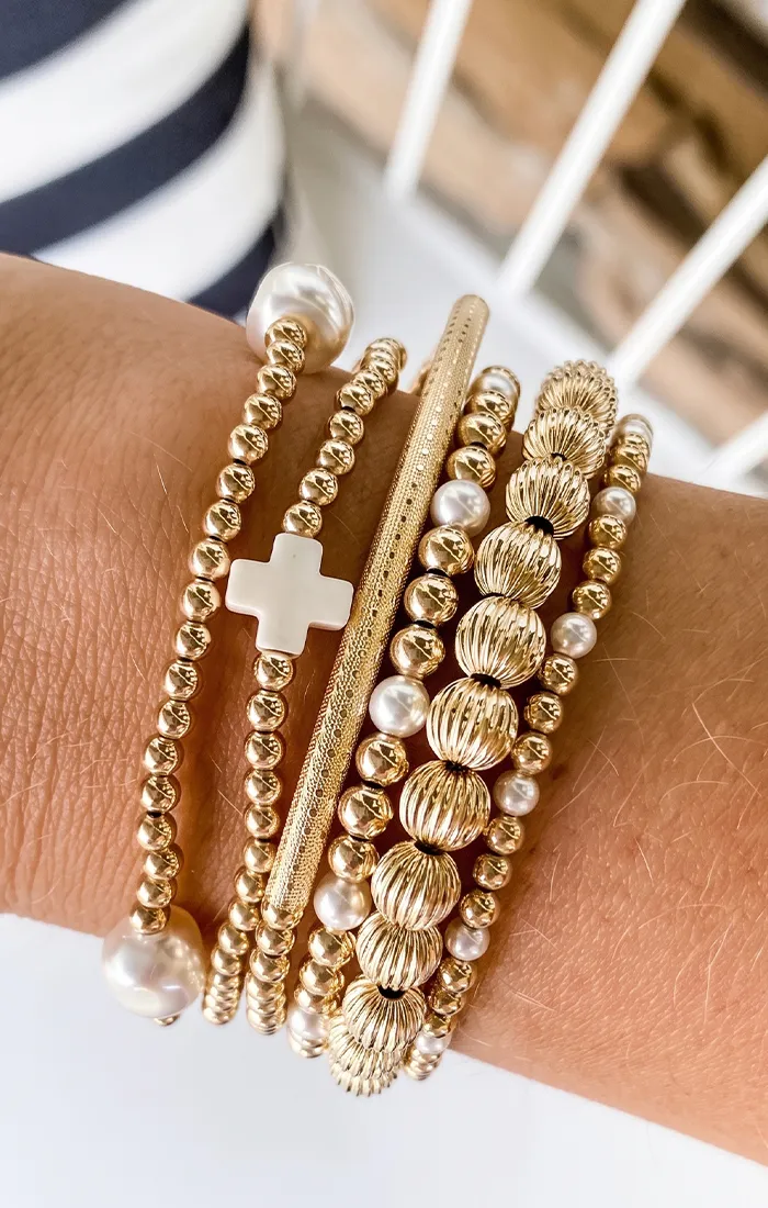 A wrist adorned with multiple gold and white beaded bracelets, featuring a cross charm.