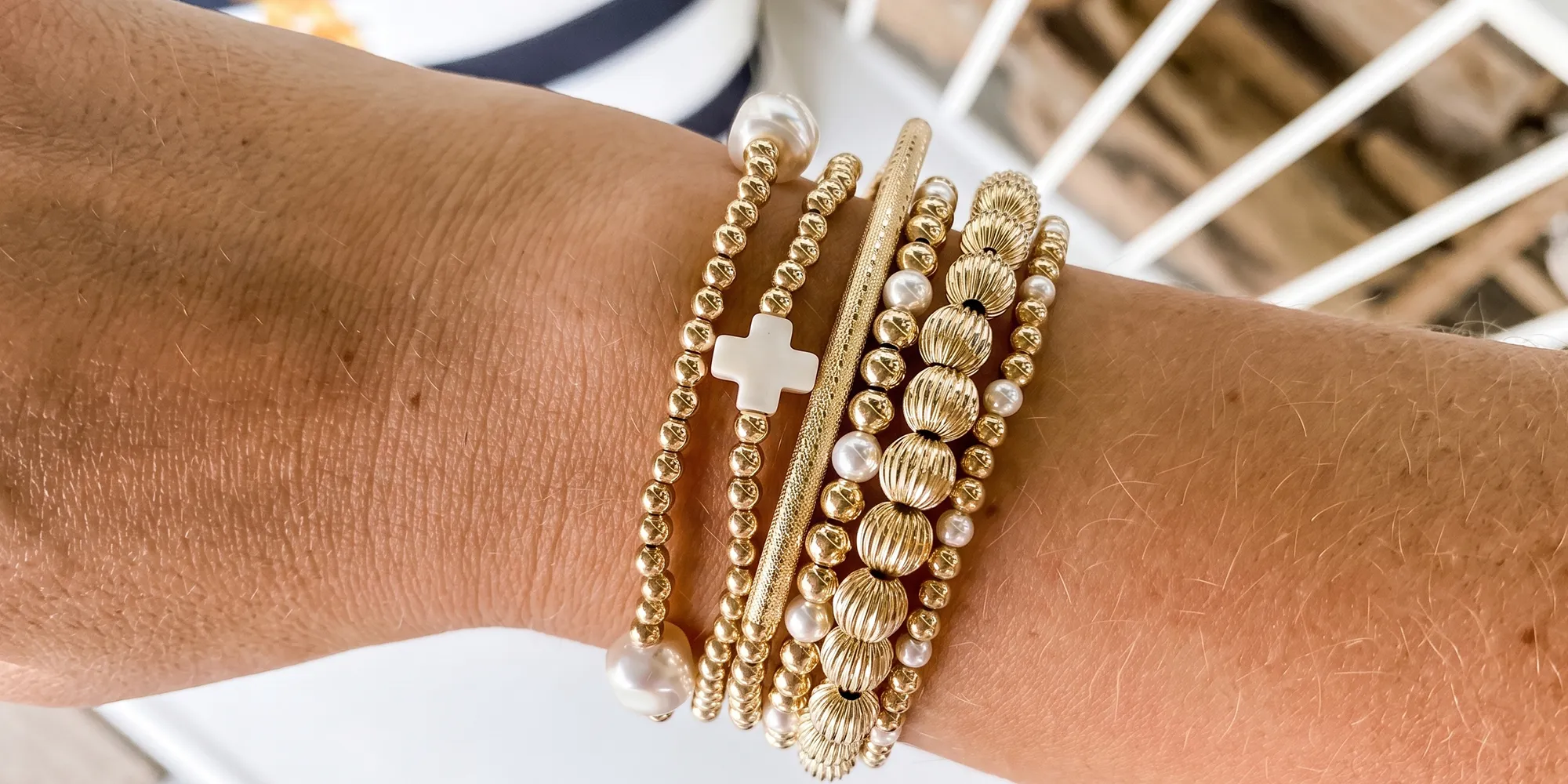 A wrist adorned with multiple gold and white beaded bracelets, featuring a cross charm.