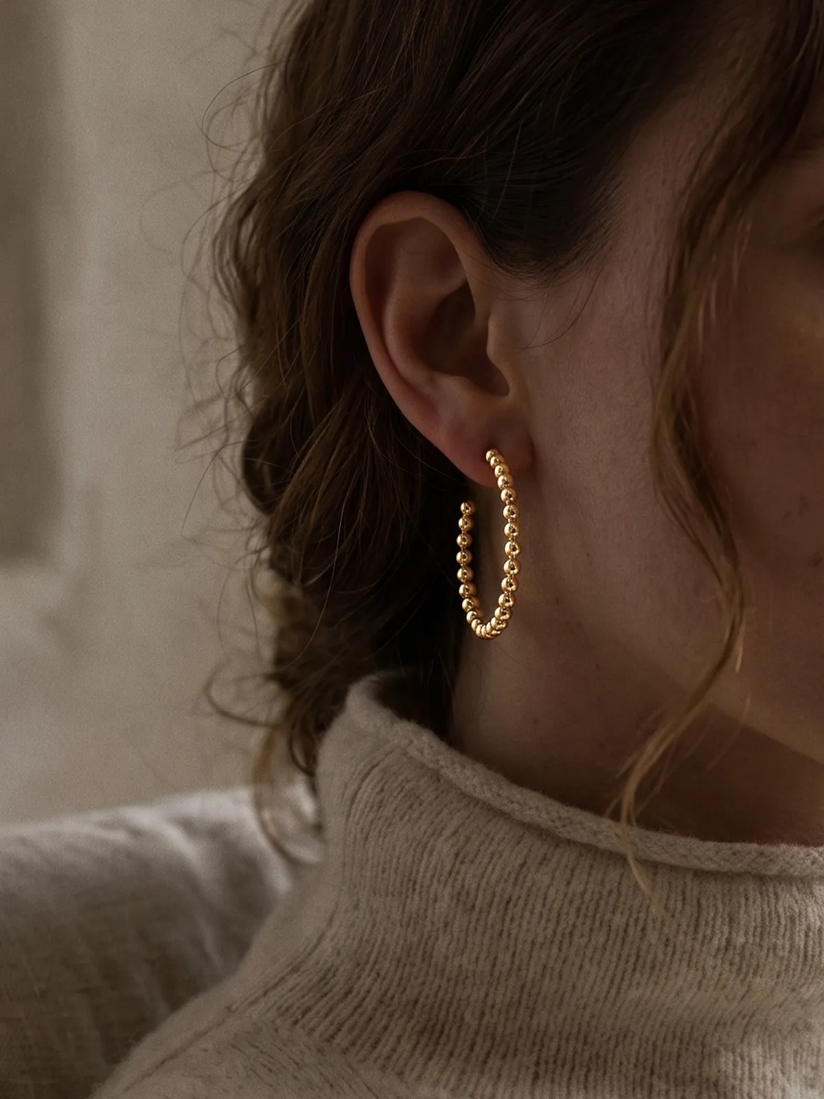 A woman wearing a gold beaded hoop earring and a beige turtleneck sweater.