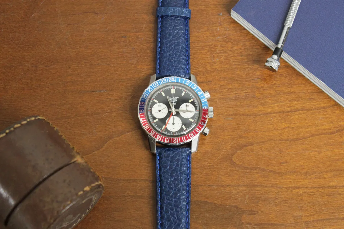 Heuer Autavia GMT Wrist watch with blue and red dial, and blue leather strap sitting on wood table