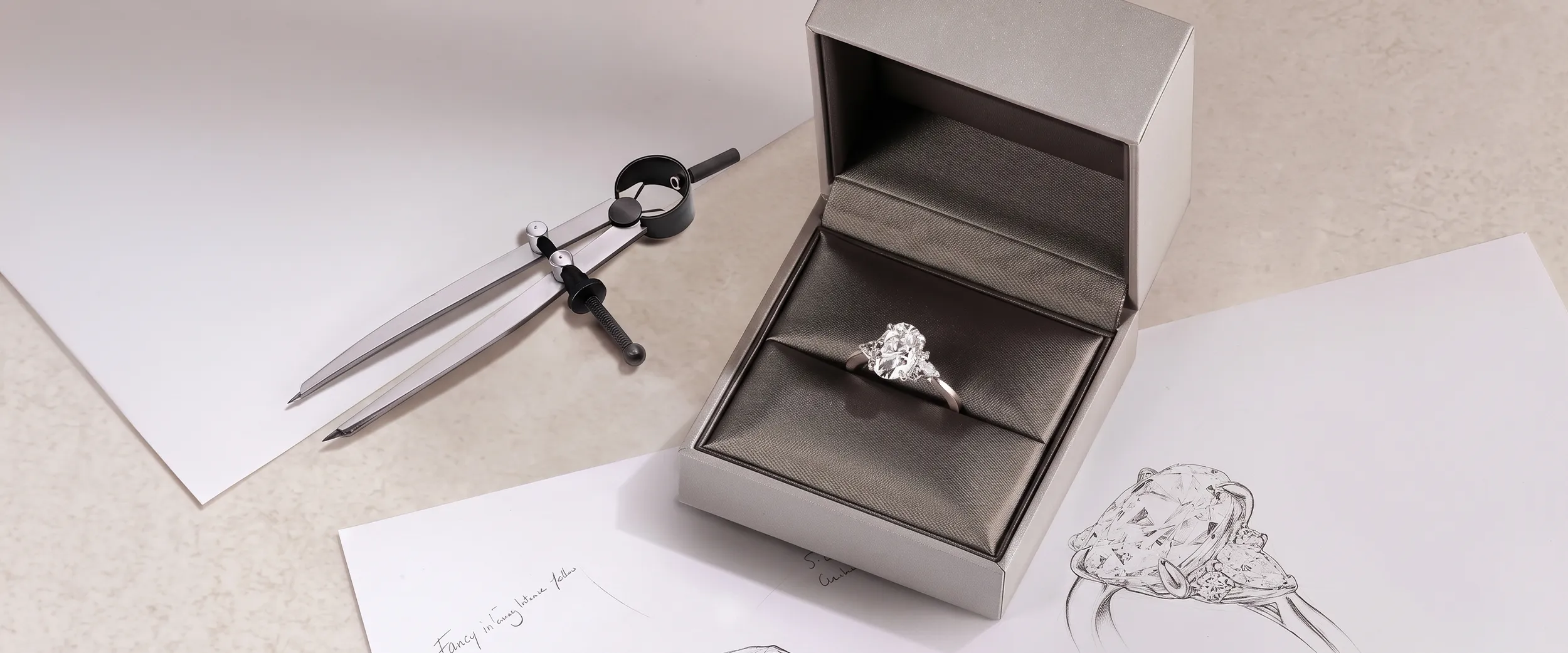 A diamond ring in a box with design sketches and a drafting compass nearby.