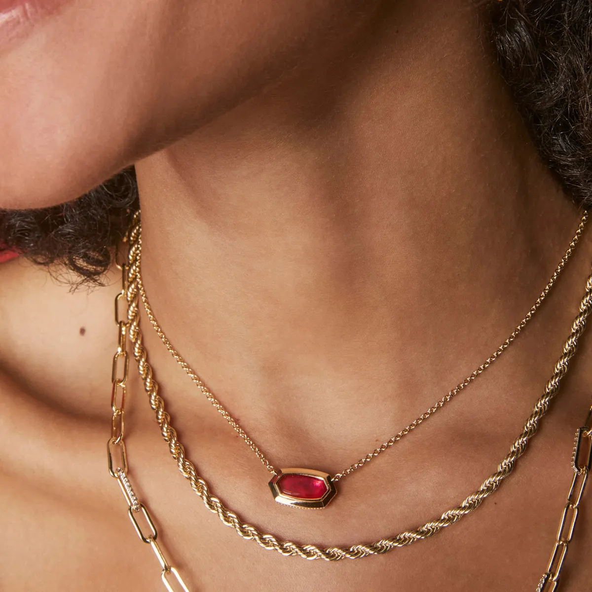 A person wearing layered gold necklaces, one with a red gemstone pendant.