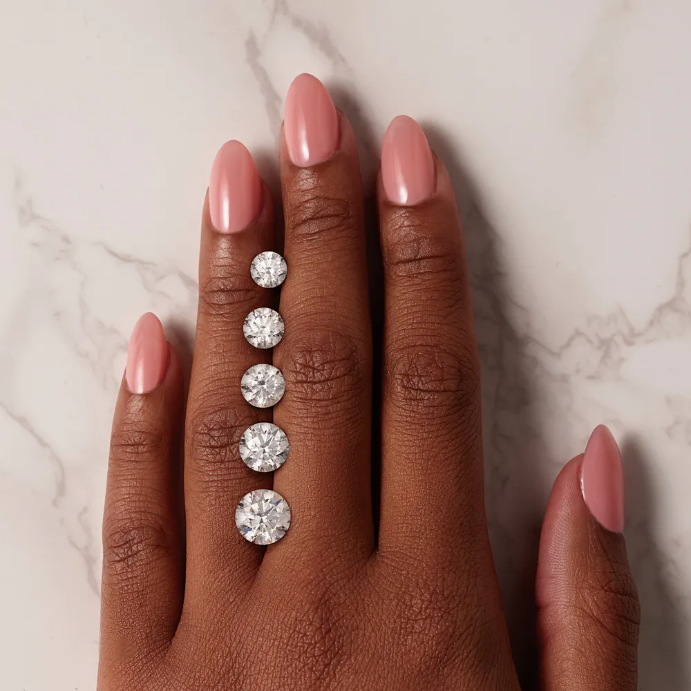 Hand with pink nails displaying five loose diamonds on a marble surface.