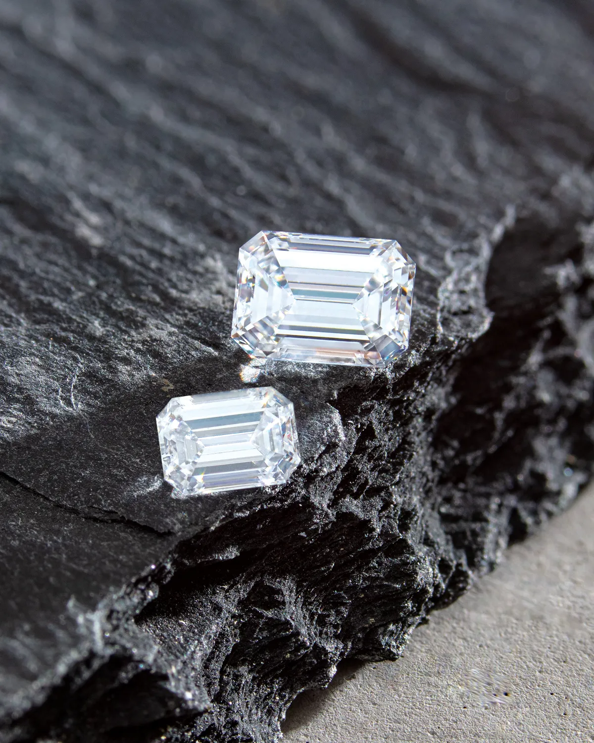 Two emerald-cut diamonds on a textured dark stone surface.