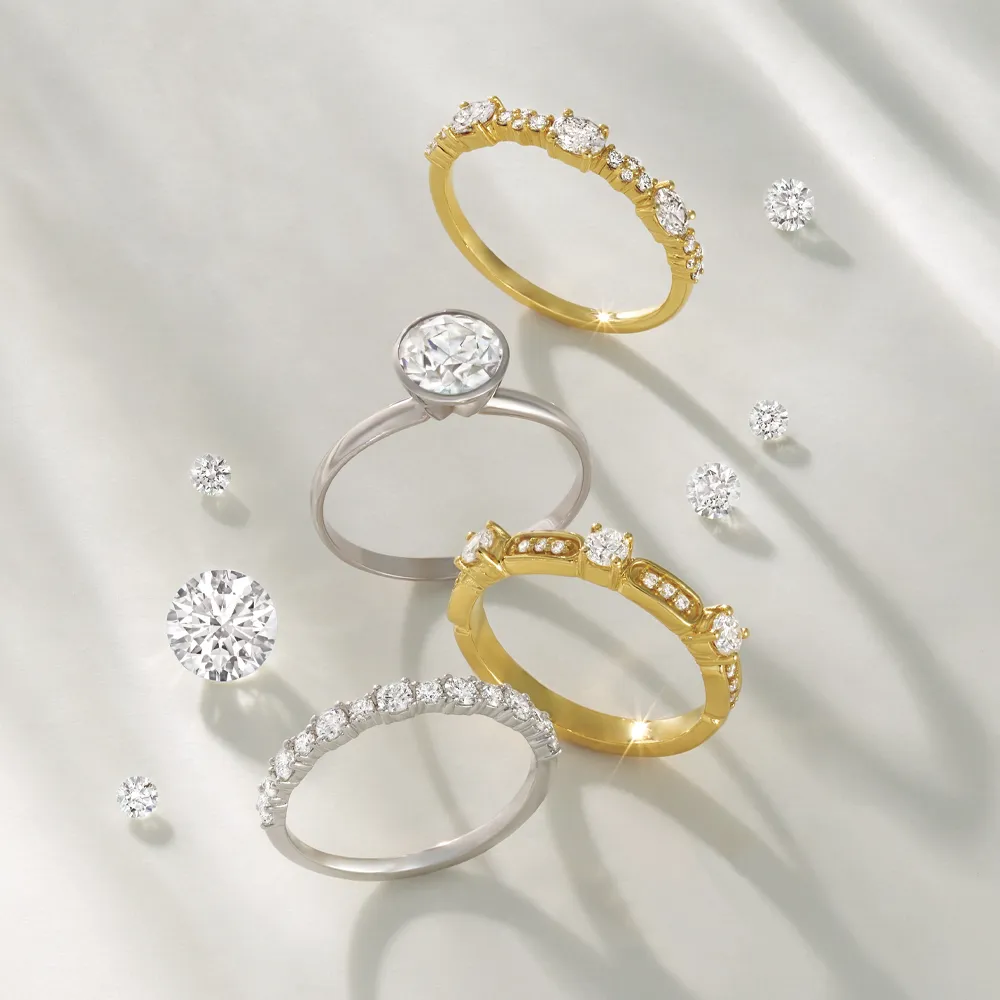 Four elegant diamond rings in gold and silver displayed with loose diamonds on a soft background.