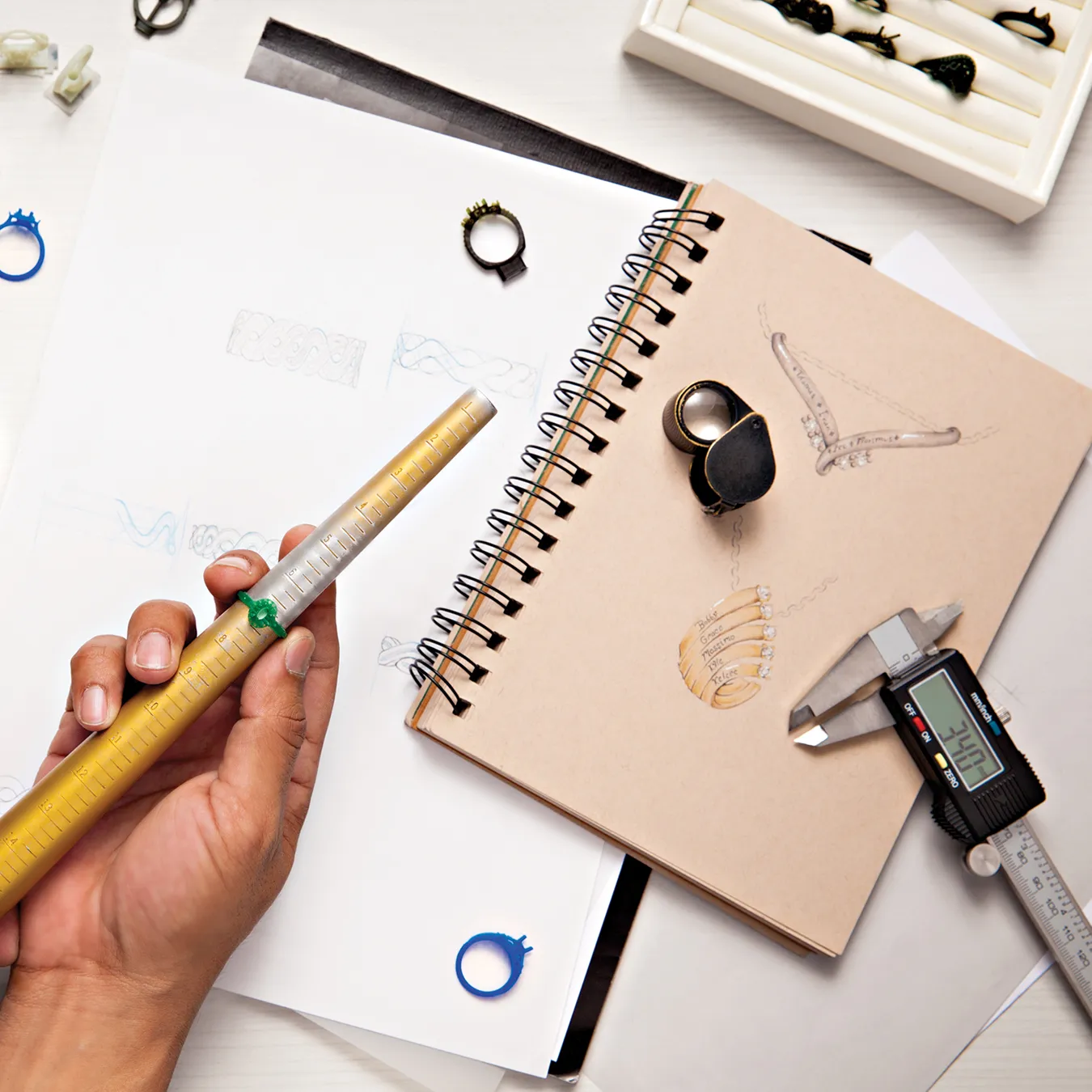 Jewelry design workspace with sketches, tools, and ring sizer on a table.