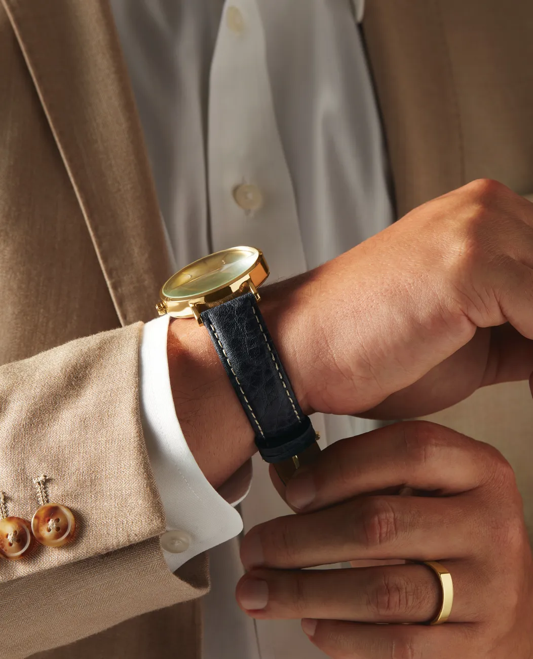 Man in beige suit adjusts gold watch with black strap.