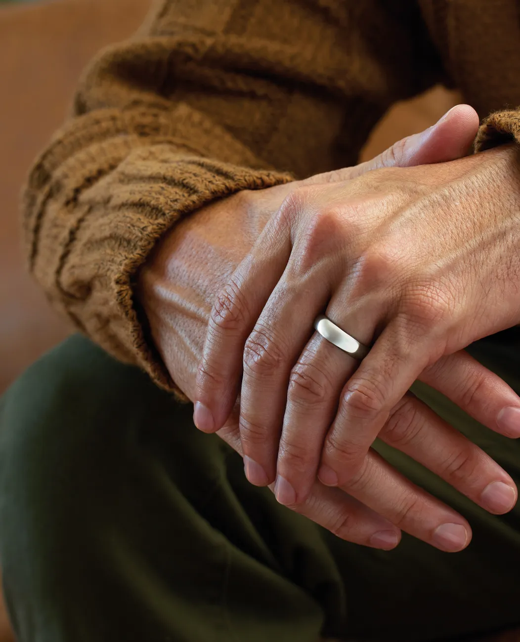 Hands with a silver wedding band, brown sweater, and green pants.