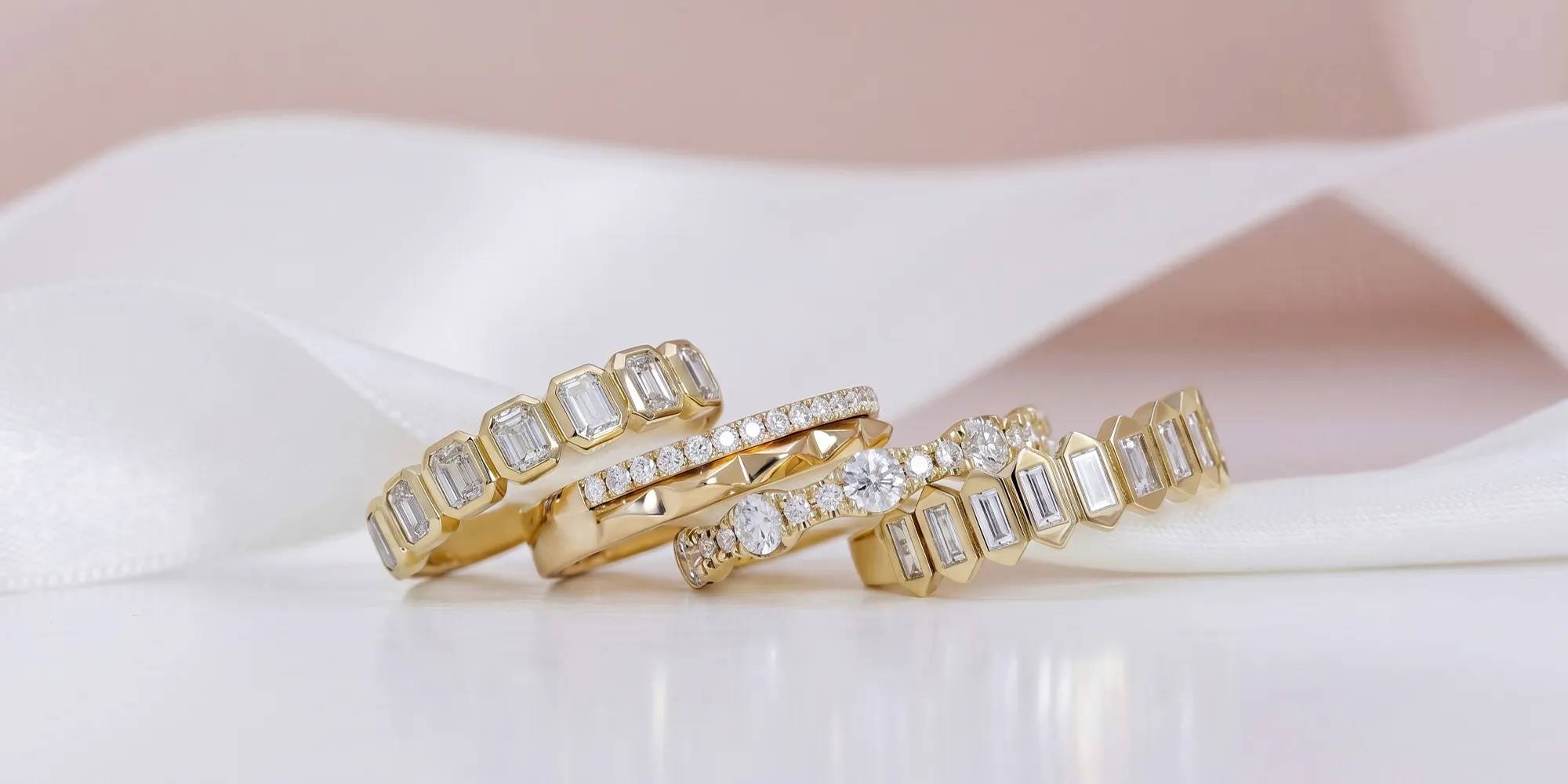 Gold rings with various diamond cuts displayed on a white surface with a ribbon backdrop.