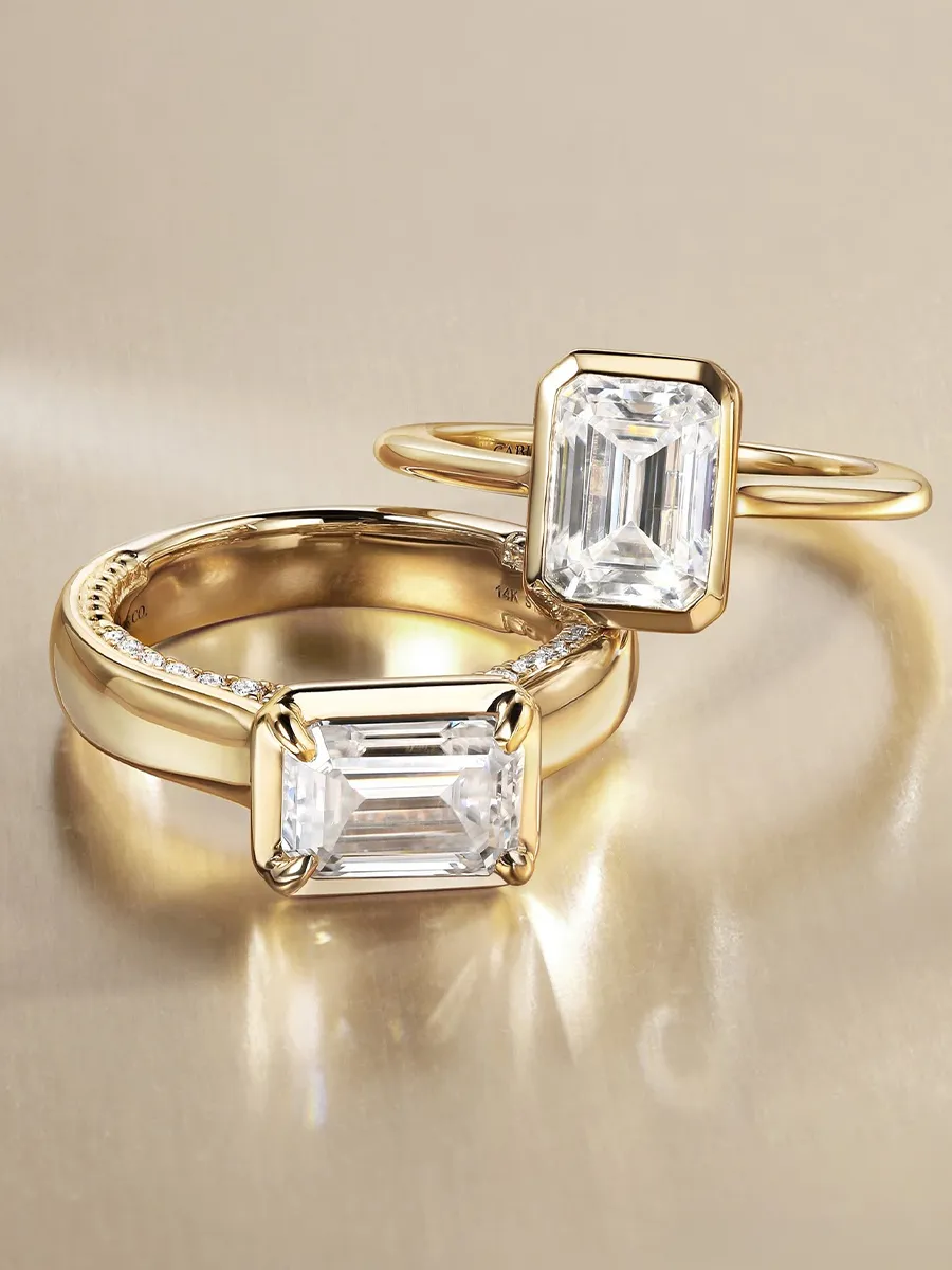 Gold rings with emerald-cut diamonds on a reflective surface.