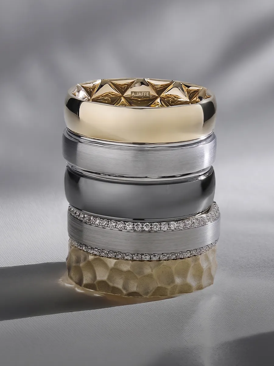 Men's wedding bands in various metals and designs stacked on a textured surface.