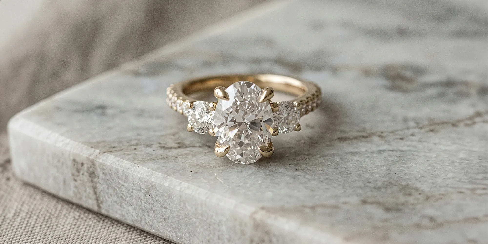 A gold engagement ring with a large oval diamond and two side stones on a marble surface.