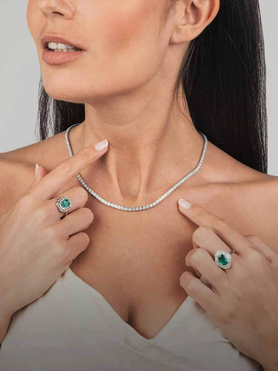 Woman showcasing a diamond necklace and emerald rings. Shop diamond necklaces at Hingham Jewelers.