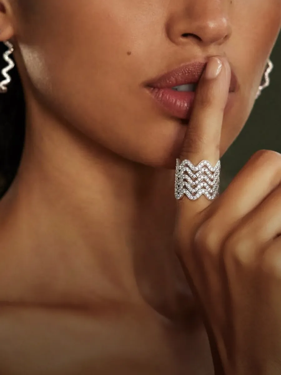 A person with a finger on lips, wearing a diamond ring and earrings.