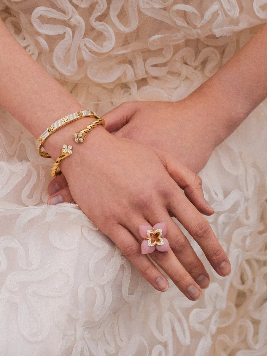Hands with gold bracelets and a pink flower ring on a textured white dress.