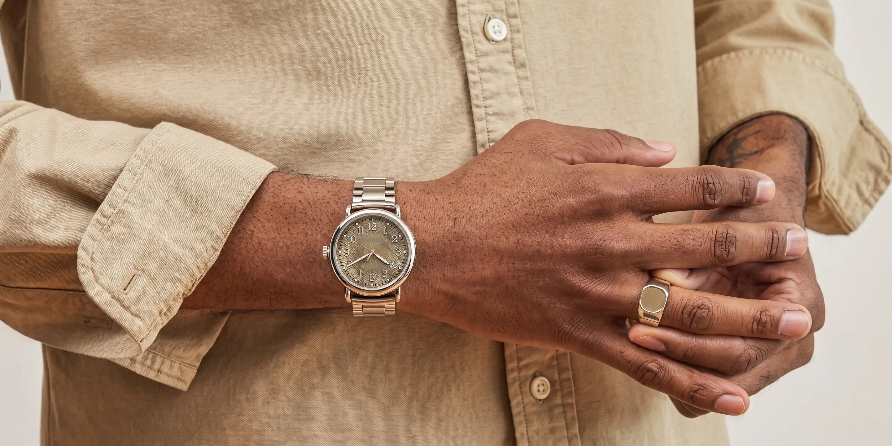 Man wearing a beige shirt, showcasing a silver watch and ring on his hands.
