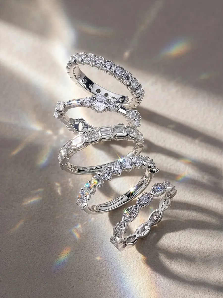 Five silver rings with various diamond settings arranged vertically on a textured surface with light reflections.