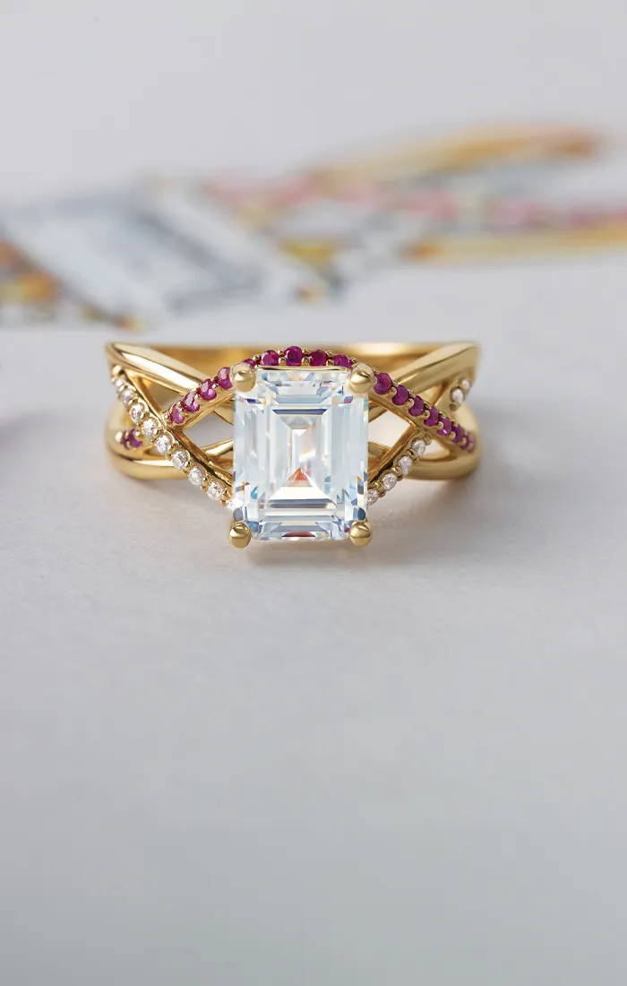 A gold ring with a large emerald-cut diamond, accented by red and white stones, on a light background.