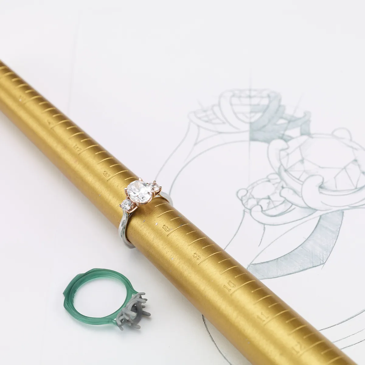 A diamond ring on a gold ring sizer with a sketch and green wax mold nearby.
