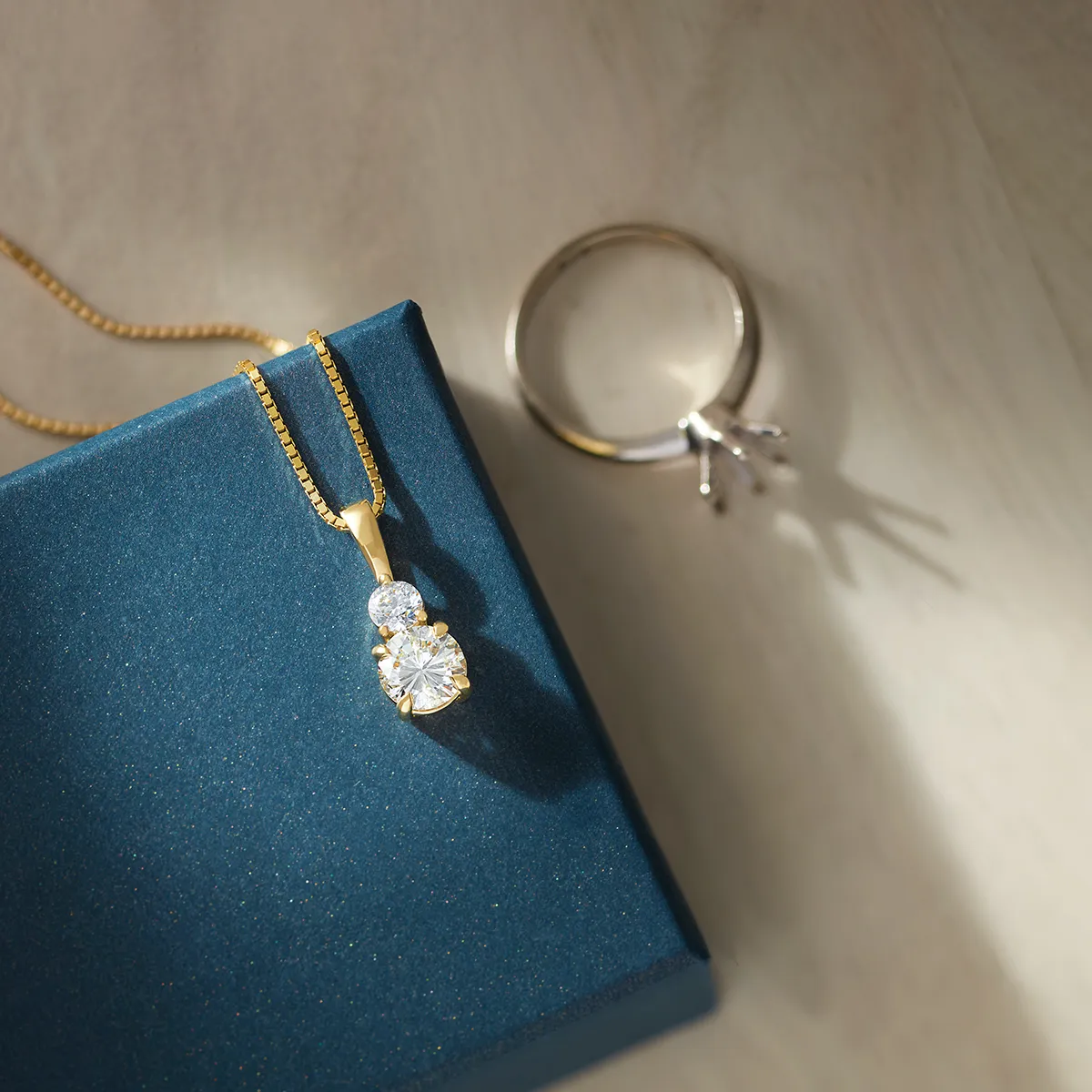 A gold necklace with a diamond pendant on a blue box, next to a ring setting.
