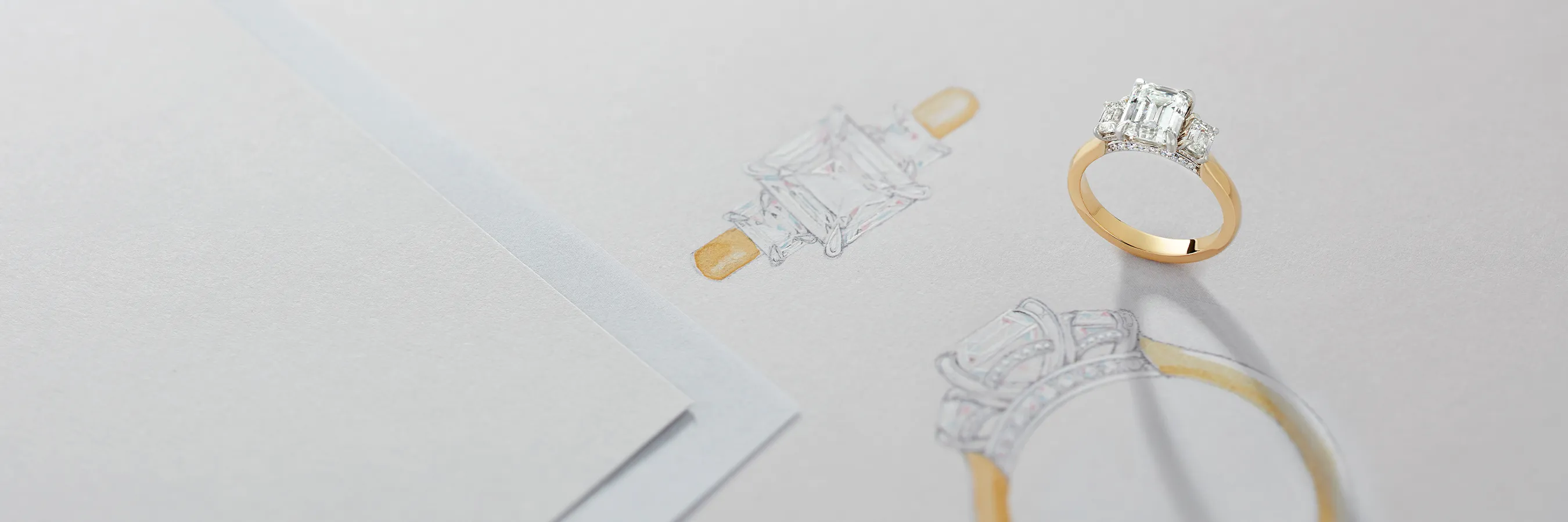 A gold ring with a large emerald-cut diamond is placed on a sketch of its design.