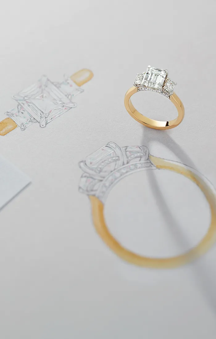 A gold ring with a large emerald-cut diamond is placed on a sketch of its design.