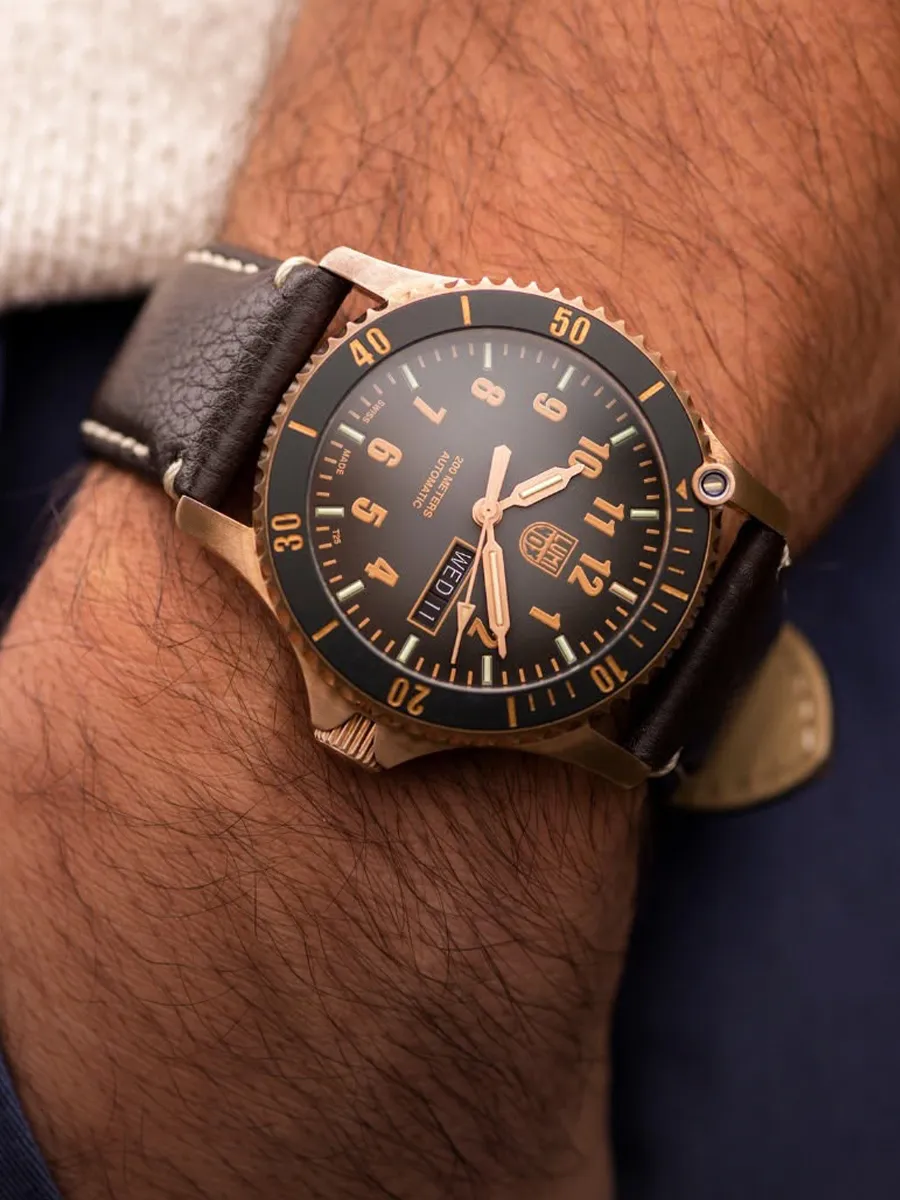 A bronze watch with a black dial and brown leather strap on a wrist.