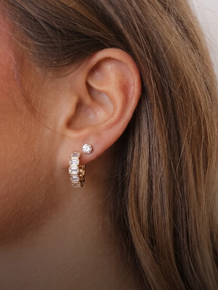 Close-up of an ear wearing a small diamond stud and a gold hoop earring with rectangular stones.