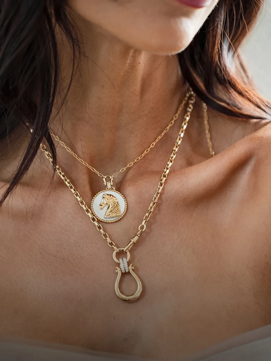 A woman wearing layered gold necklaces, one with a horse pendant and another with a hook-shaped charm.
