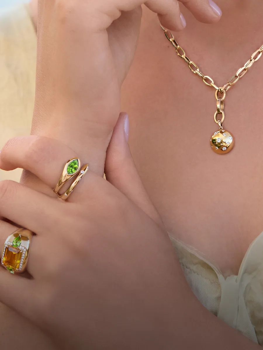 A person wearing a gold necklace with a pendant and two rings with green and yellow gemstones.