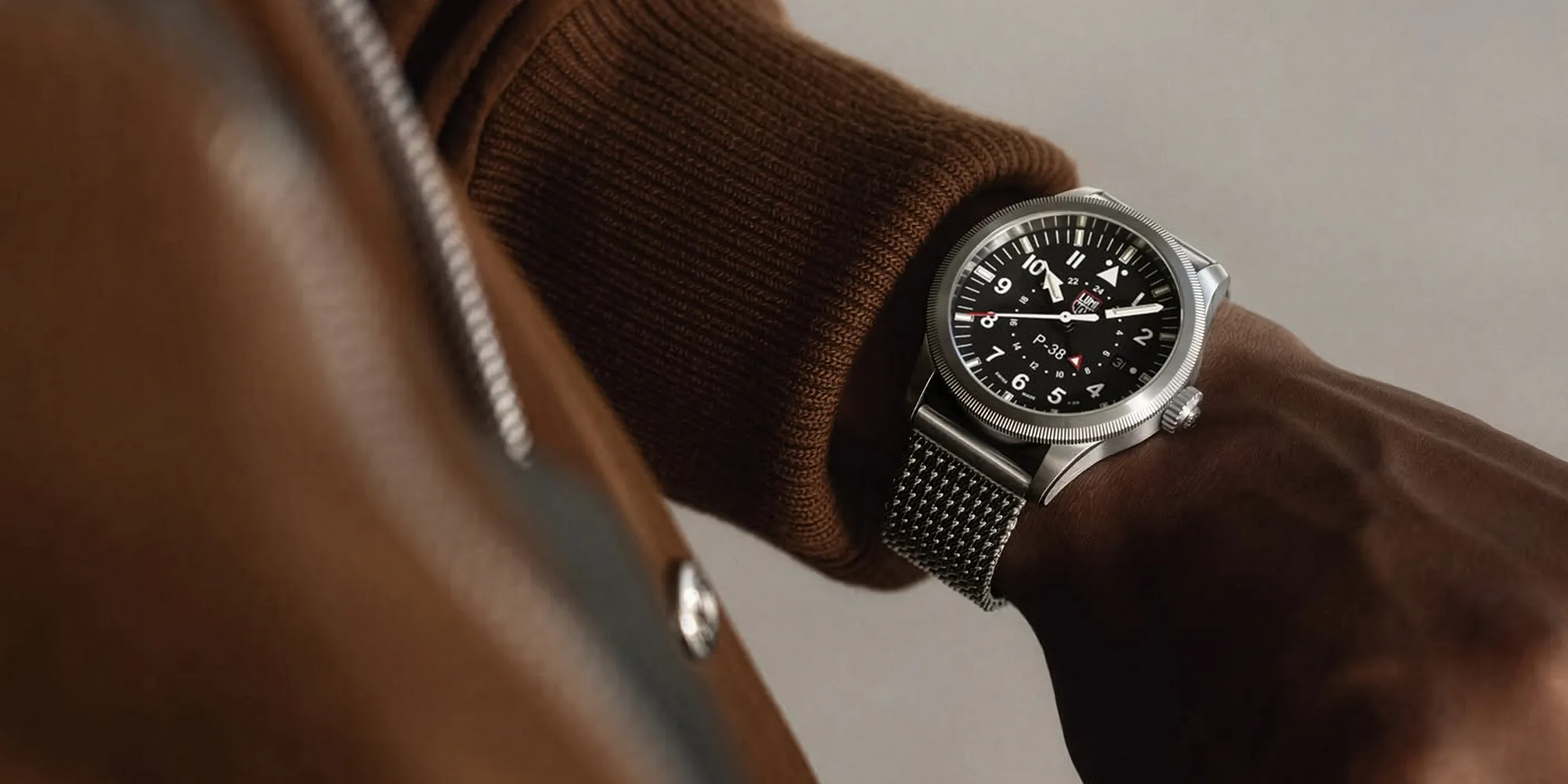 A person wearing a brown jacket and a silver watch with a black dial and mesh strap.