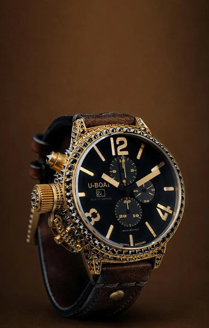 Luxurious gold and black watch with a studded bezel and brown leather strap on a brown background.