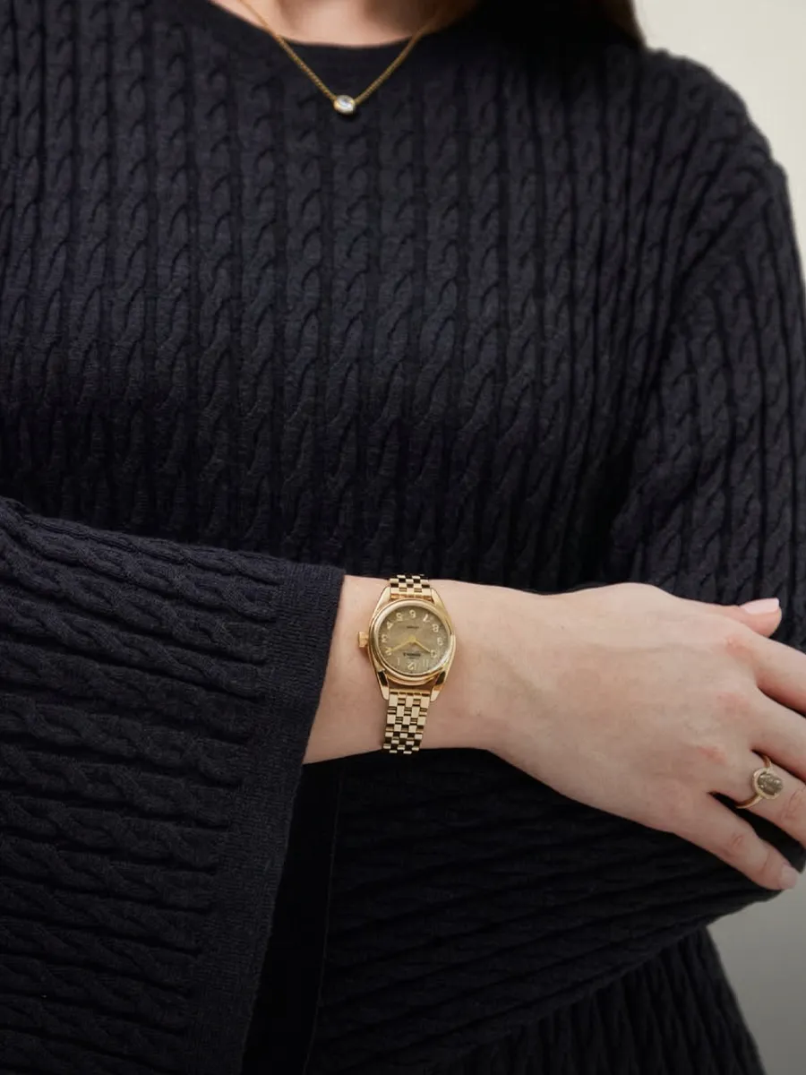 A person wearing a gold watch and a black cable-knit sweater, with a necklace and ring.