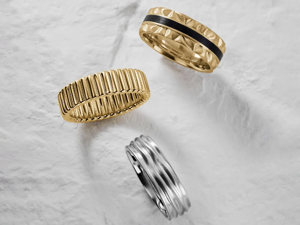 Three men's wedding bands: one gold with ridges, one silver with grooves, and one gold with a black inlay.