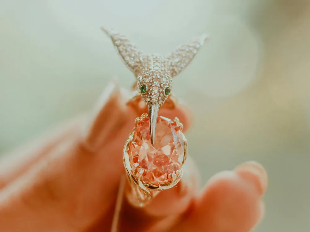 A sparkling hummingbird ring with a pink gemstone, held delicately in a hand.