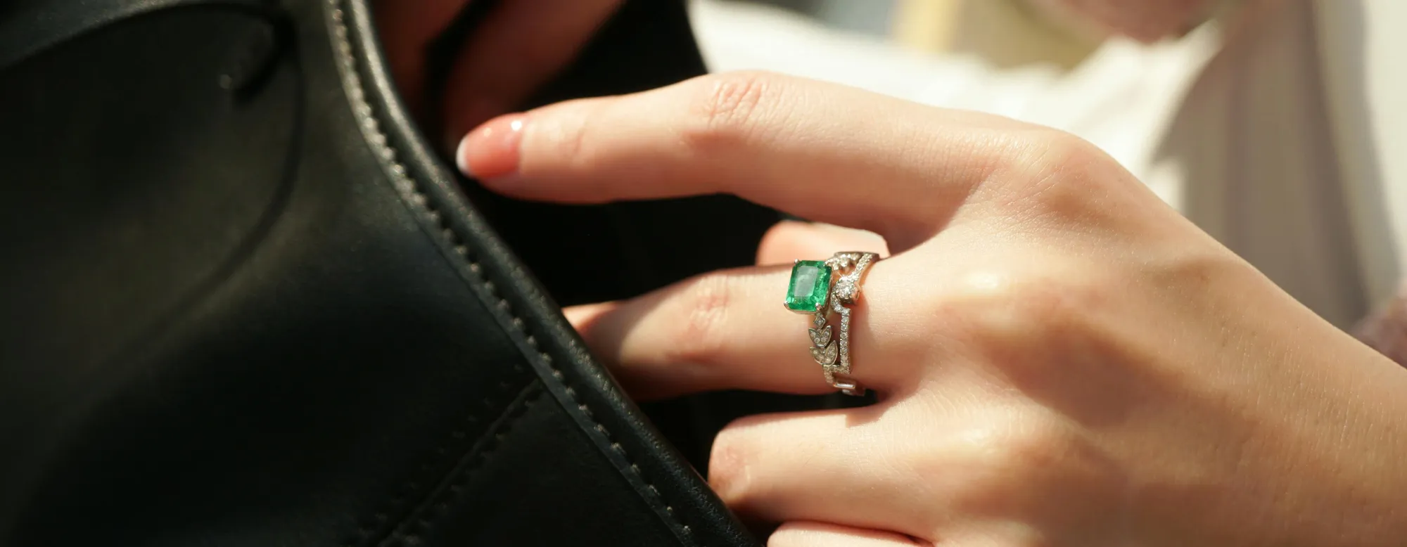 A hand with a green emerald ring touches a black leather bag, showcasing fine jewelry.
