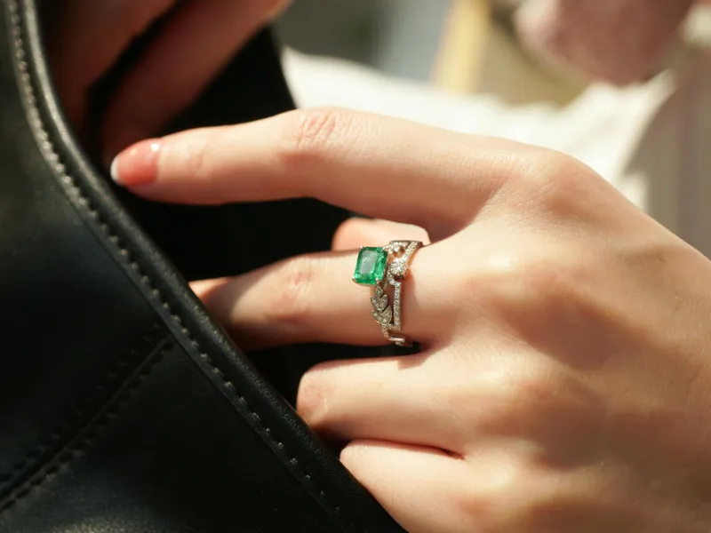 A hand with a green emerald ring touches a black leather bag, showcasing fine jewelry.