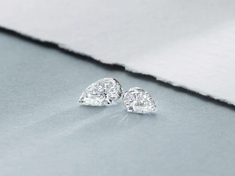 Two sparkling loose diamonds in pear and round shapes on a soft gray surface.