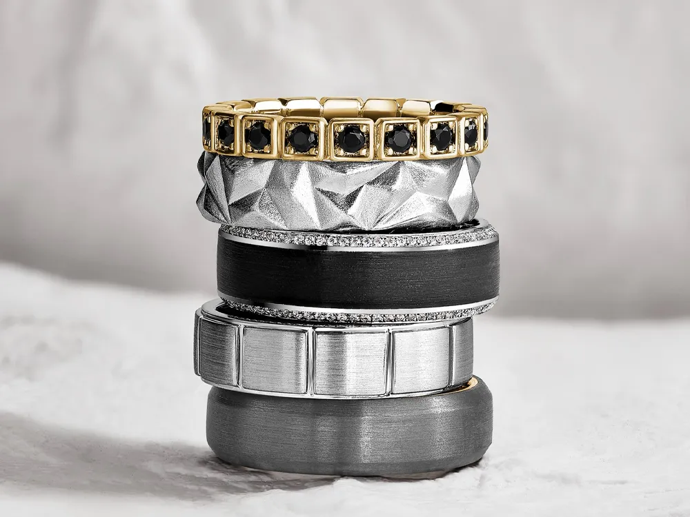 Stack of elegant men's wedding bands in various metals and designs, showcasing fine jewelry.