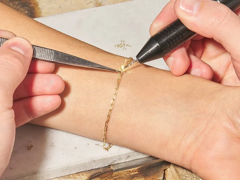 A person using tweezers and a pen to attach a gold chain bracelet on a wrist, showcasing permanent jewelry.