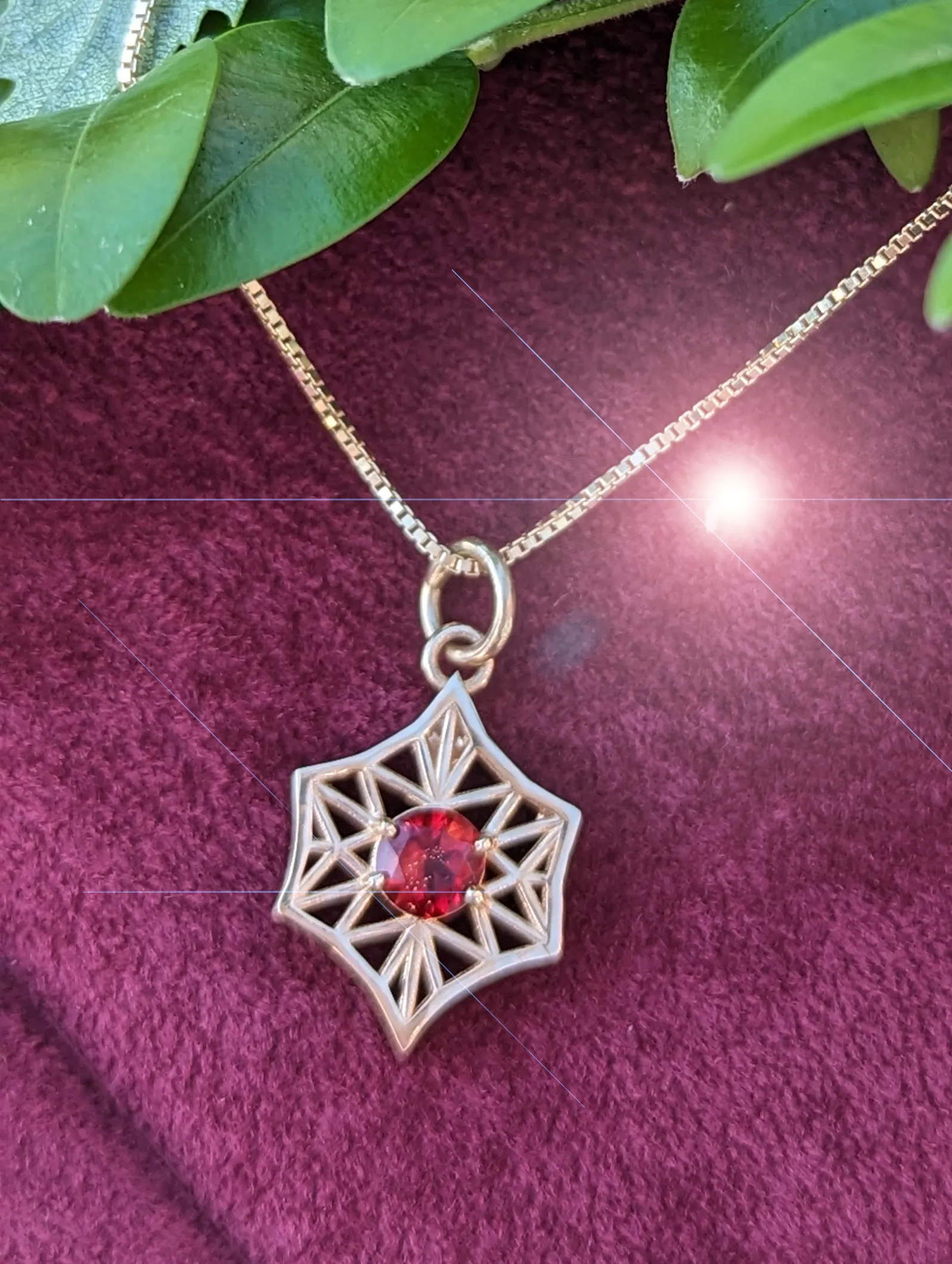 A star-shaped pendant with a red gemstone on a gold chain, set against a purple background with green leaves.