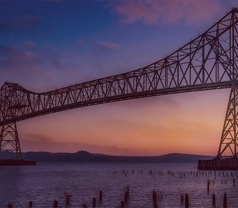 Astoria-Megler Bridge at sunset over water, with silhouettes of pilings in the foreground. Click to learn about our store histor
