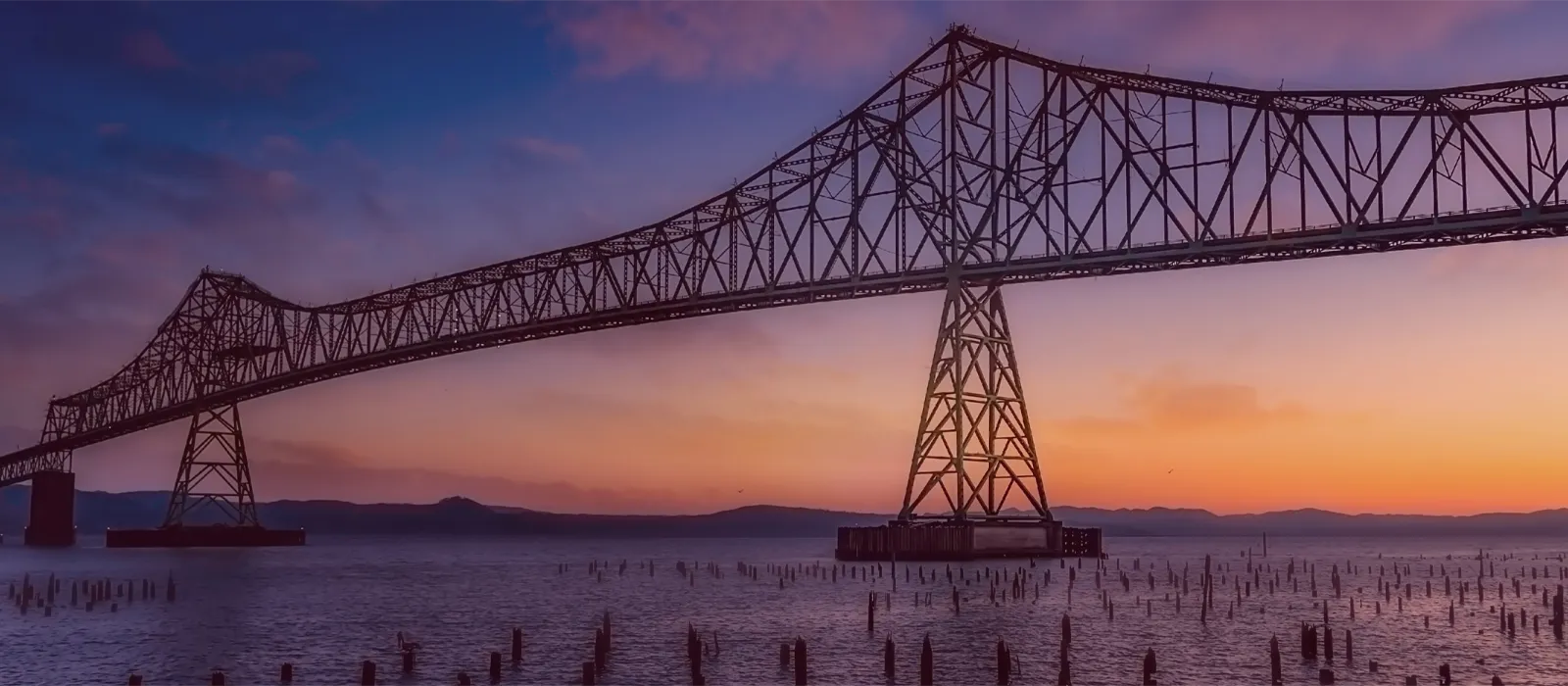 Astoria-Megler Bridge at sunset over water, with silhouettes of pilings in the foreground. Click to learn about our store histor