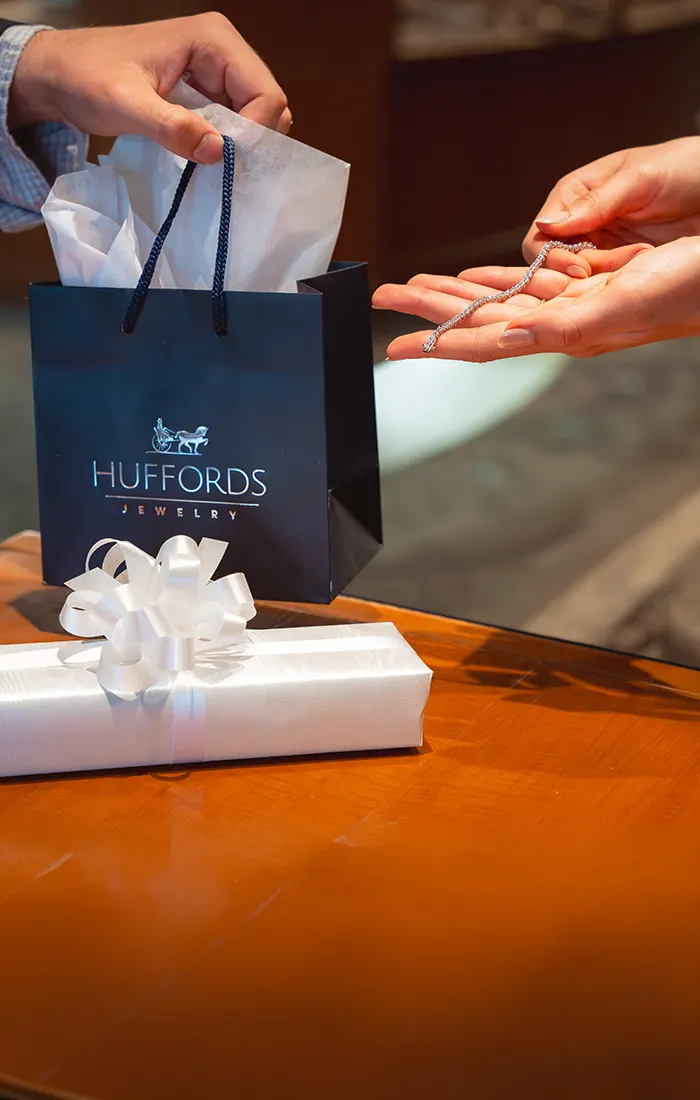 Hands holding a diamond bracelet over a Huffords Jewelry gift bag. Click to contact us.